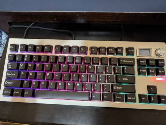 A tenkeyless mechanical keyboard (specifically the Zuoya GMK87) on a table, featuring a white case, white on black keycaps, and on the right side, a small screen and a volume knob.