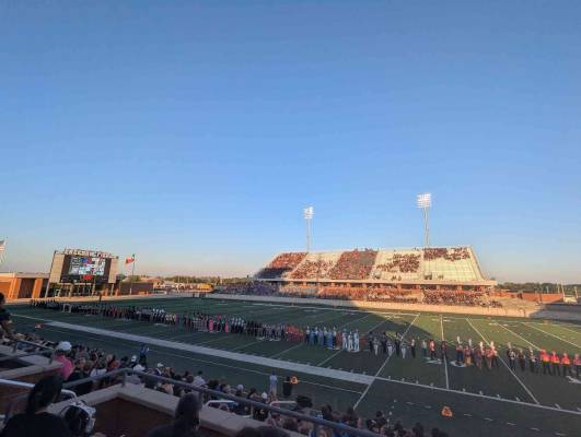 A football field showing drum majors from various high school bands in a straight line that takes up nearly the whole field. Sitting in the stands on the opposite side from my perspective is the band members from the many marching bands that competed today.